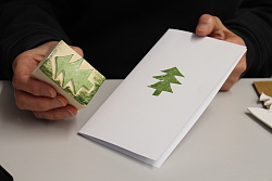 Man sieht auf der linken Bildhälfte ein rechteckiges Holzstück, das schräg in die Kamera gehalten wird. Darauf ist ein grün eingefärbtes Stück Moosgummi in Form einer Tanne zu sehen. Rechts im Bild wurde das Motiv des Tannen-Stempels auf einen weiße Karte gedruckt.