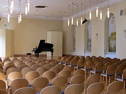 Das Bild zeigt den Pflugsaal im Haus zum Pflug. Es sind mehrere Stuhlreihen zu sehen. Der Blick geht Richtug Bühne, auf der ein schwarzer Flügel steht. Der Raum hat hellgelbe Wände und einen hellen Parkettfußboden.