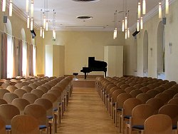 Das Bild zeigt den Pflugsaal in Haus zum Pflug. Der Blick geht über die Stuhlreihen im Zuschauerraum in Richtung Bühne. Dort steht ein schwarz glänzender Flügel. Der Raum wirk mit seinen hellgelben Wänden, dem hellen Parkettboden und den großen Fenstern einladend und freundlich. 